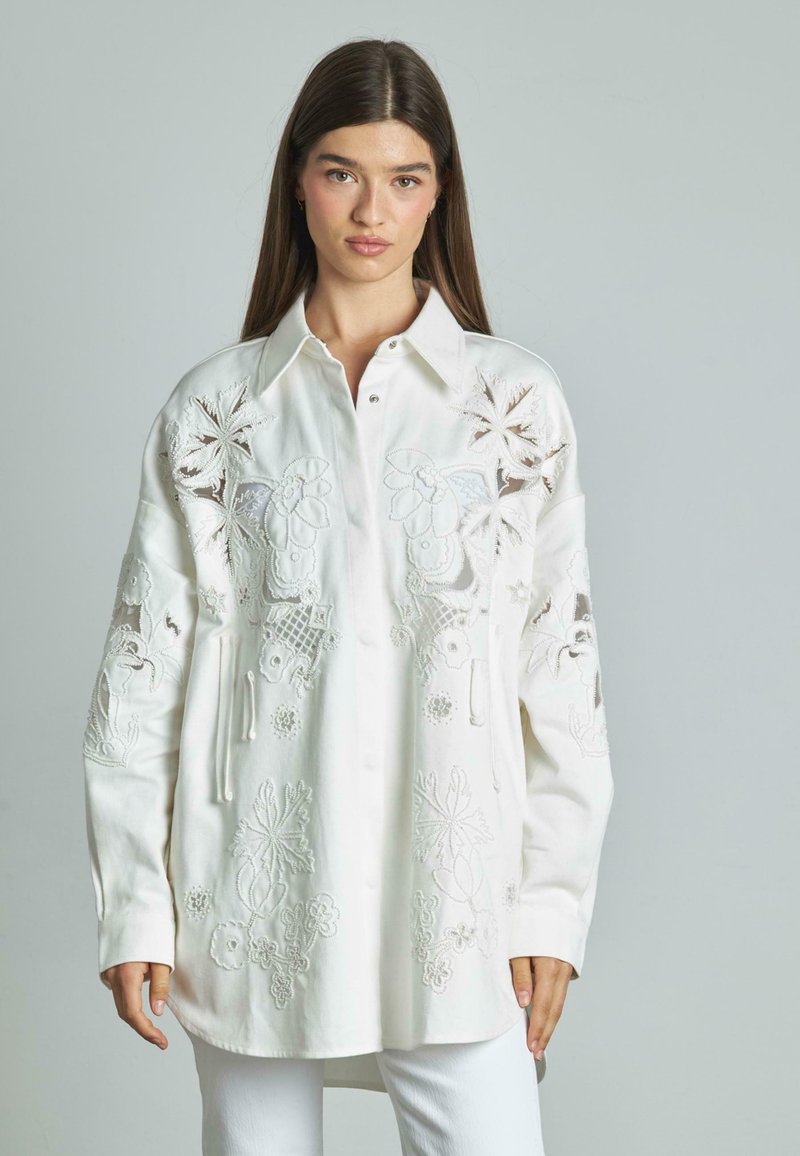 Camisa blanca bordada con diseños florales y de hojas, con cuello y cierre de botones en la parte delantera y mangas largas. Textura de tela suave.
