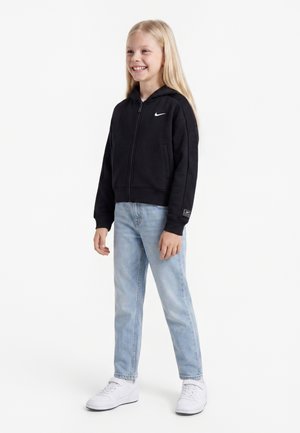 Enfant souriant aux longs cheveux blonds portant un sweat à capuche noir Nike zippé, un jean bleu clair et des baskets blanches, debout devant un fond uni.