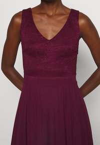 Burgundské šaty s krajkovým korzetem a hlubokým výstřihem ve tvaru V, mají hladkou, splývavou sukni. Texturovaná krajka a široký detail pasu.