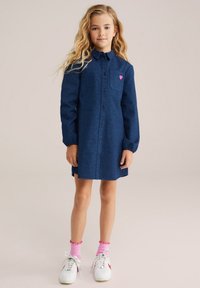 Abito blu navy con bottoni, motivo a cuori, maniche lunghe e un accento a forma di cuore rosa sul petto. Indossato con scarpe da ginnastica bianche e calze rosa.