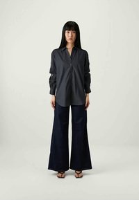 Camisa escura de botões com mangas longas e detalhe de enrolar, combinada com calças largas em azul-marinho, usada com chinelos sobre um fundo simples.
