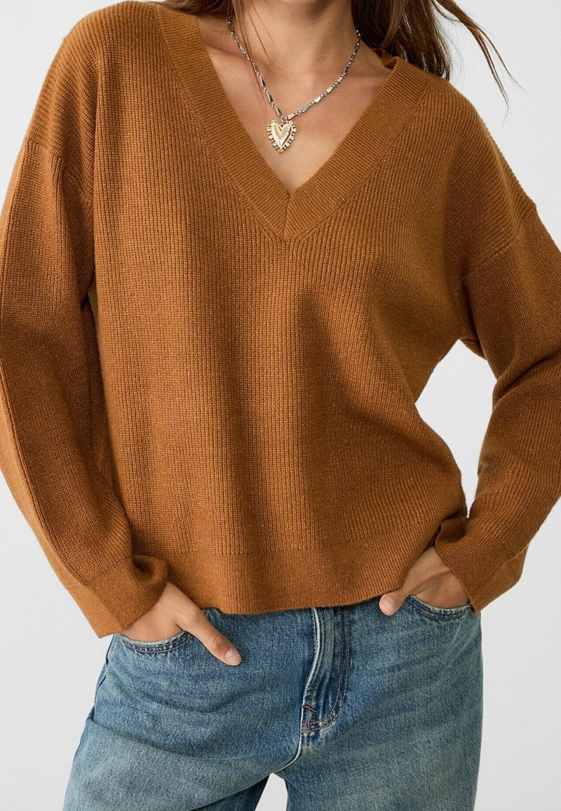 Femme portant un pull en tricot marron à col en V et un jean bleu, les mains dans les poches, avec un collier pendentif argenté en forme de cœur.
