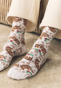 Calzini lavorati a maglia con sfondo beige e motivo leopardato, decorati con biscotti marroni e foglie di agrifoglio verdi.