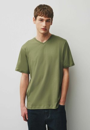 Bărbat tânăr cu păr castaniu scurt, purtând un tricou cu decolteu în V verde măsliniu și blugi închiși la culoare, stând în fața unui perete simplu, de culoare deschisă.