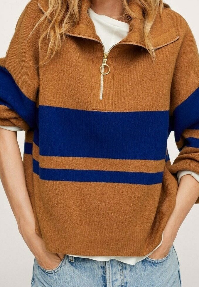 Personne portant un pull en maille marron avec des rayures horizontales bleues et un col demi-fermeture éclair, les mains dans les poches d'un jean en denim bleu clair.