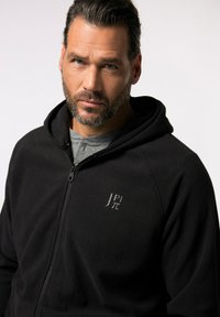 Zwarte fleece ritssluiting hoodie met een logo op de linker borst, voorzien van een capuchon en een gladde textuur, gedragen over een grijs shirt.