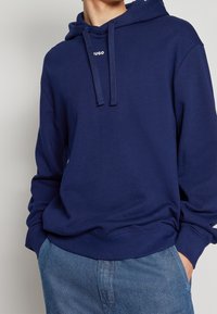 Námořnická modrá mikina s kapucí, stahovací šňůrkou, klokánkovou kapsou, žebrovanými manžetami a lemem. Na přední straně má decentní logo detail. Měkká textilie.