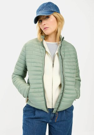 Veste verte matelassée avec un devant zippé, un col montant et des poches latérales. Portée sur une chemise rayée blanche et marine, associée à un jean bleu.
