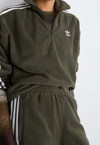 Olivgrön fleece-pullover med en kvart dragkedja, med en vit logotyp och randiga detaljer på ärmarna, tillsammans med matchande joggers.