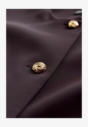 Mørk lilla stof med en glat tekstur, prydet med en gylden spiralknap som en fremtrædende detalje. Stoffet hænger blødt.