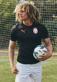 Maglietta atletica nera a maniche corte con logo ed emblema arancione, pantaloni corti bianchi, mentre tiene in mano un pallone da calcio multicolore su un campo d'erba.