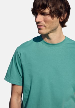 Mladý muž s krátkými hnědými vlasy, který má na sobě modrozelené krátké bavlněné tričko, hledí stranou proti jednobarevnému pozadí.