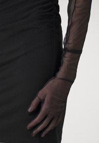 Svart tajt klänning med rynkade detaljer, kombinerad med genomskinliga svarta nät-handskar med vågiga kanter. Texturerad tyg förstärker det visuella djupet.