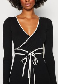 Robe portefeuille noire côtelée avec manches longues, bordure blanche, décolleté en V profond et détail noué à la taille. Texture lisse et silhouette ajustée.