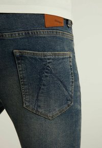 Jean en denim avec un lavage foncé, présentant une étiquette en cuir marron, des coutures visibles et un design distinctif de poche arrière angulaire.