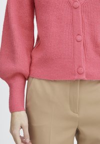 Cardigan rosa de canalé con un acabado texturizado, que presenta tres botones redondos en la parte delantera y mangas ajustadas, combinado con pantalones beige.