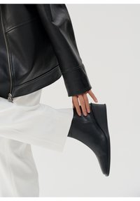 Černá kožená kotníková bota s elegantním designem, s hladkým povrchem, zaoblenou špičkou a nízkým klínovým podpatkem. Obklopena bílými kalhotami a bundou.