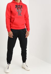 Person som bär en röd hoodie med texten "JUST DO IT.", svarta joggingsbyxor och vita sneakers, stående mot en vit vägg.