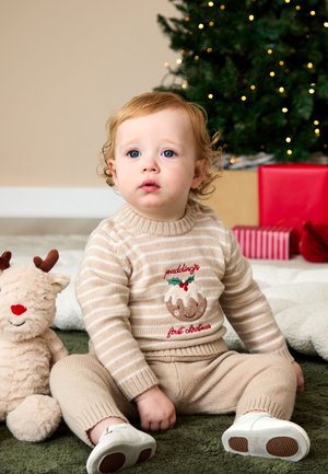 Dojčenské dieťa so kučeravými vlasmi v béžovom vianočnom svetri a nohaviciach sedí na zelenom koberci blízko plyšového soba, sviatočného stromčeka a zabalených darčekov.