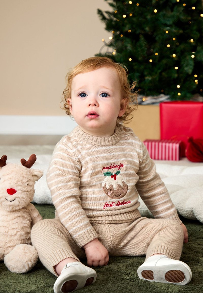 Bambino piccolo con capelli ricci indossa un maglione di Natale beige e pantaloni, seduto su un tappeto verde vicino a una renna di peluche, un albero di Natale e regali confezionati.