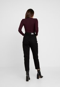 Svarta höga byxor och en ribbad mörkröd topp, sedd bakifrån. Toppen har långa ärmar och byxorna har en etikett på baksidan.