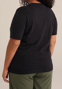 Svart stickad kortärmad topp med strukturerat prickmönster, ribbad halsringning och fåll. Bärs med gröna byxor och visar en avslappnad passform.