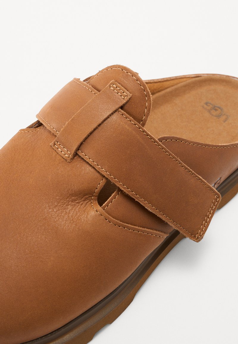 Braune Leder-Slipper mit einem breiten Kreuzriemen, sichtbaren Nähten und einer gepolsterten Innensohle. Abgerundete Zehenpartie und flexible Sohle.