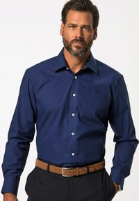 Námořnická modrá košile s dlouhým rukávem a límečkem na knoflíky, přední kapsou a bílými knoflíky; hladká textura; nošena s hnědým opaskem a tmavými kalhotami.