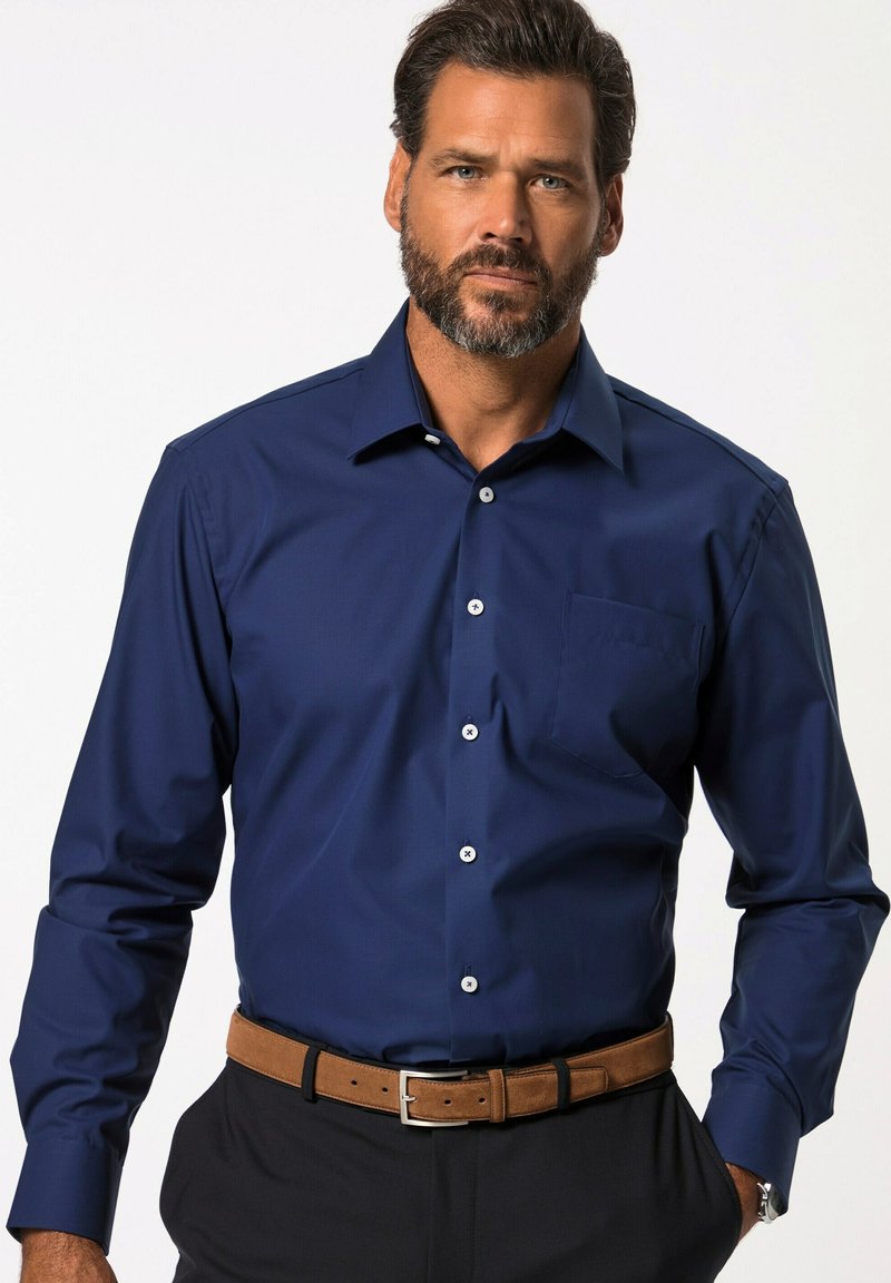 Námořnická modrá košile s dlouhým rukávem a límečkem na knoflíky, přední kapsou a bílými knoflíky; hladká textura; nošena s hnědým opaskem a tmavými kalhotami.