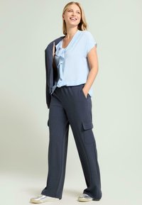 Lyseblå, flæsekantet bluse med korte ærmer kombineret med mørkeblå cargo-bukser med sidelommer. Modellen har metallic sneakers på.