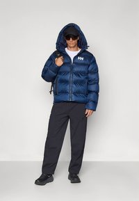 Marineblaue Steppjacke mit horizontalen Nähten, Reißverschluss vorne und verstellbarer Kapuze. Kombiniert mit schwarzer Hose und Sportschuhen, getragen mit einer Sonnenbrille.