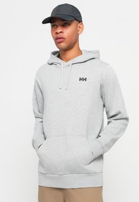 Sivá mikina s kapucňou a predným vreckom, so šnúrkou na kapucni a čiernym logom na hrudi. Má mäkkú tkaninu a voľný strih.