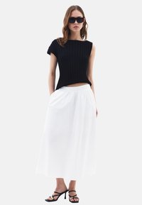 Top nero a coste con maniche corte, gonna bianca fluida e sandali neri con tacco alto e dettagli dorati. La modella indossa occhiali da sole.