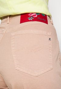 Vaatteet vaaleanpunaiset farkut punaisella brodeeratulla kukkakuviolla, joka on vyötärövyön kohdalla. Huomionarvoista taka-taskun ompeluaihetta.