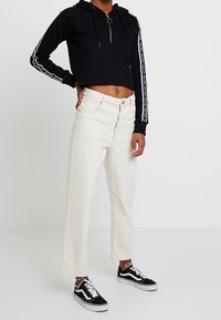 Černá zkrácená mikina s předním zipem a kroužkovým táhlem, kombinovaná s volnými bílými džíny a černými teniskami s bílými akcenty.