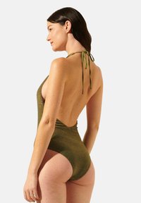 Maillot de bain une pièce vert olive avec un dos échancré, des liens tour de cou et un tissu brillant texturé. Les ouvertures de jambe allongées rehaussent la silhouette.