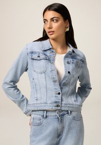 Veste en denim bleu clair au design ajusté, dotée de deux poches poitrine, de quincaillerie en argent et d'accentuations texturées sur l'ensemble.