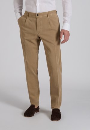 Uomo che indossa pantaloni beige plissettati, camicia bianca con bottoni e mocassini marrone scuro, in piedi con le mani nelle tasche davanti a uno sfondo semplice.