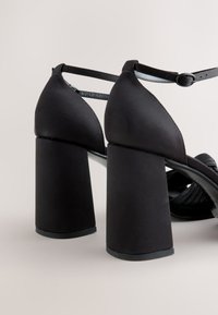 Fekete magas sarkú szandálok selyemfényű felülettel, széles tömzsi sarkakkal és vékony bokapántokkal. A dizájn letisztult, a lábujjrész finoman ráncolt.