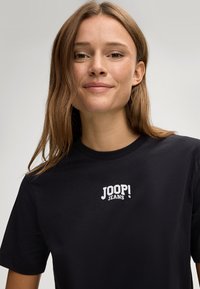 Schwarzes Baumwoll-T-Shirt mit einem weißen Logo "JOOP! JEANS" auf der Brust, kurzen Ärmeln und rundem Ausschnitt. Glatte Textur.