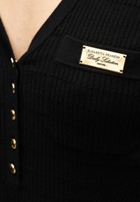 Μαύρο ριμπ ύφασμα ένδυμα με χρυσά κουμπιά και χρυσή ορθογώνια ετικέτα που  γράφει "Elisabetta Franchi Daily Seduction".