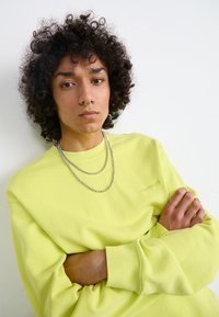 Neon gul sweatshirt med rund halsudskæring og ribbede manchetter, to sølvkæder lagdelt omkring halsen, model med krøllet hår og armene crossed.