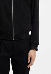 Black suede bomber jacket with a zipper, ribbed cuffs and hem, featuring side pockets and a smooth texture. Black trousers visible underneath.