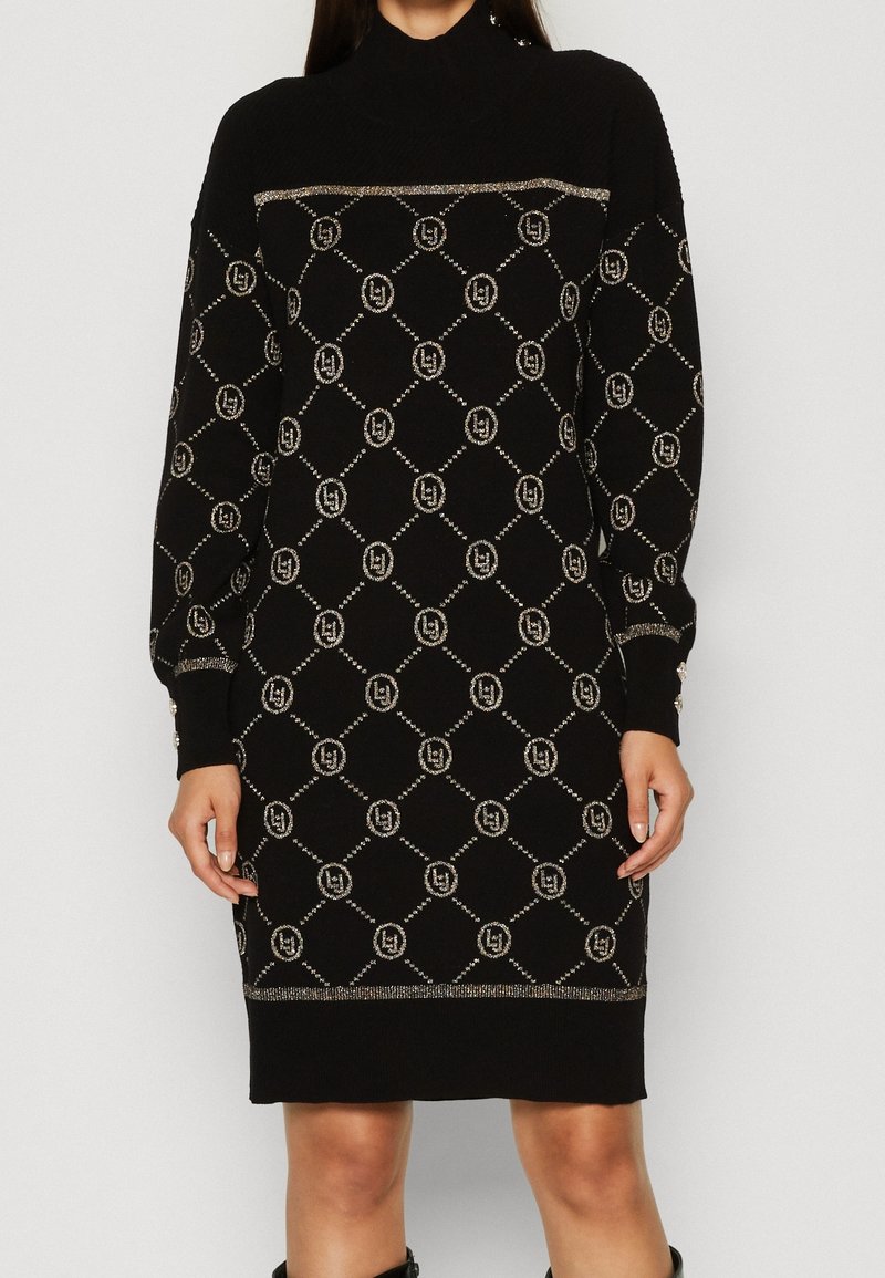 Robe en tricot noire avec col montant, présentant un motif en diamant doré associé à un design de logo. Les manches longues et les poignets texturés apportent une touche de détail.