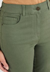 Groene katoenen broek in een mixstof met een gestructureerde afwerking. Heeft een knoopsluiting aan de voorkant, riemlussen en een zijzak. Close-up van het taillebandgebied.