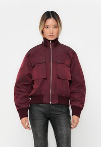 Woman wearing a burgundy bomber jacket with front flap pockets, ribbed cuffs and hem, paired with textured black pants, standing against a white background.