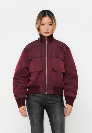 Donna che indossa una giacca bomber bordeaux con tasche frontali a pattina, polsini e orlo a coste, abbinata a pantaloni neri textured, in piedi contro uno sfondo bianco.