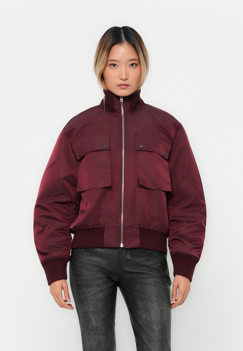 Woman wearing a burgundy bomber jacket with front flap pockets, ribbed cuffs and hem, paired with textured black pants, standing against a white background.