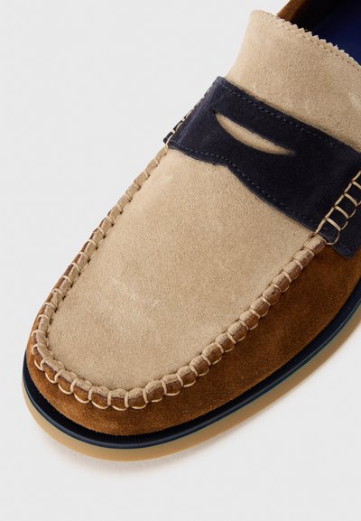 Pantof suede în stil mocasin, cu o schemă de culori bej și albastru închis, detalii cusute și o decupare pe călcâi. Textură moale și formă rotundă.