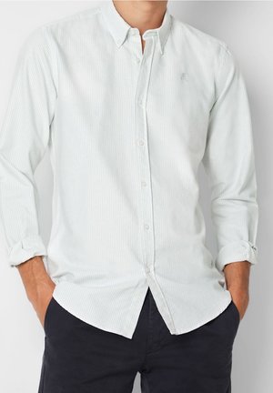 Mężczyzna w jasnoniebieskiej koszuli w paski z guzikami, z podwiniętymi rękawami i ciemnych spodniach, ręce w kieszeniach, na jednolitym tle.